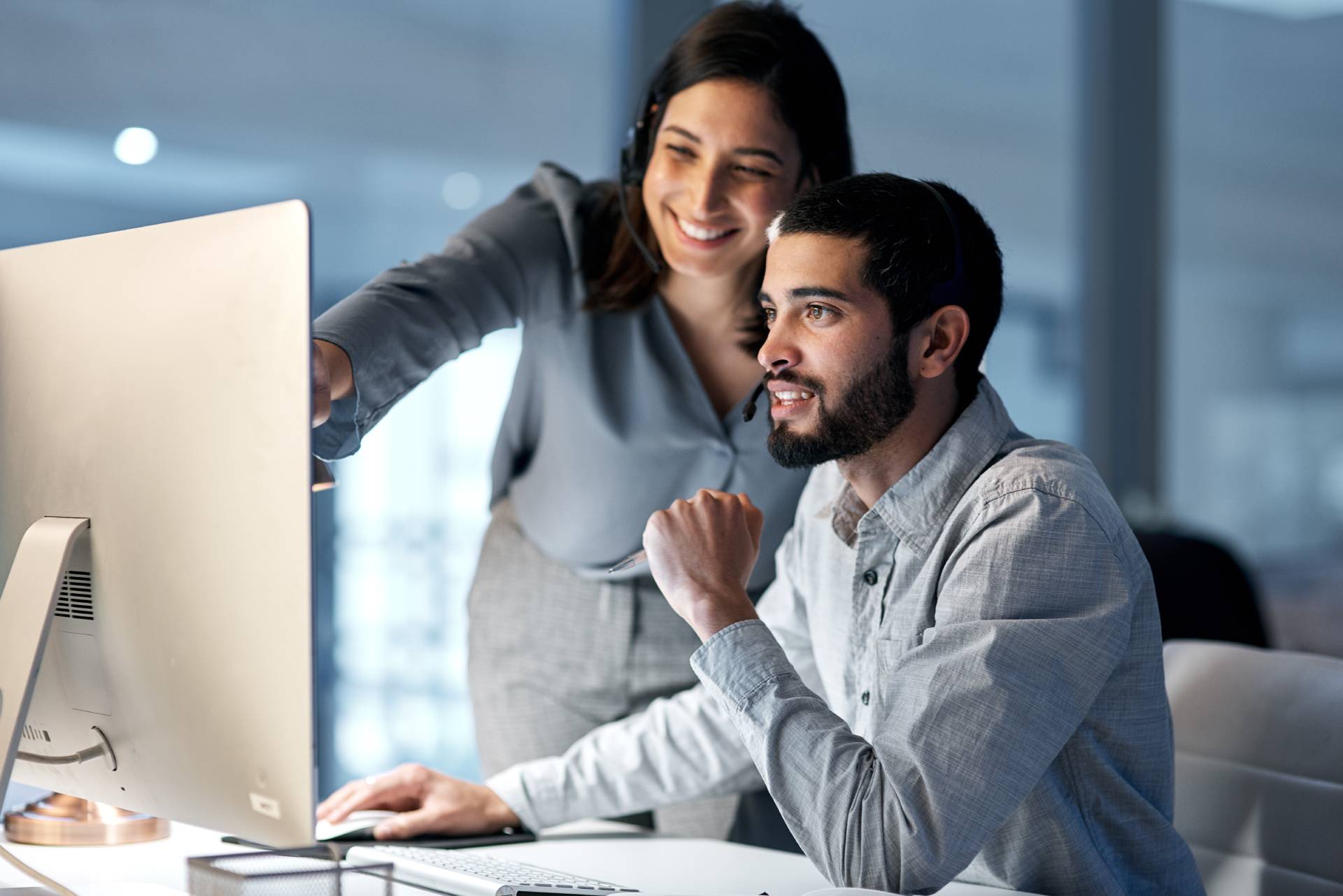 How-to-Choose-Licensing-Management-Software Two colleagues in an office smile as a woman points at a computer screen, assisting a seated man. The scene is collaborative and positive.